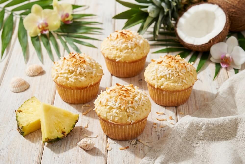 Muffinki kokosowo-ananasowe na talerzu, wokół kawałki ananasa i połówka kokosa w tropikalnej aranżacji.