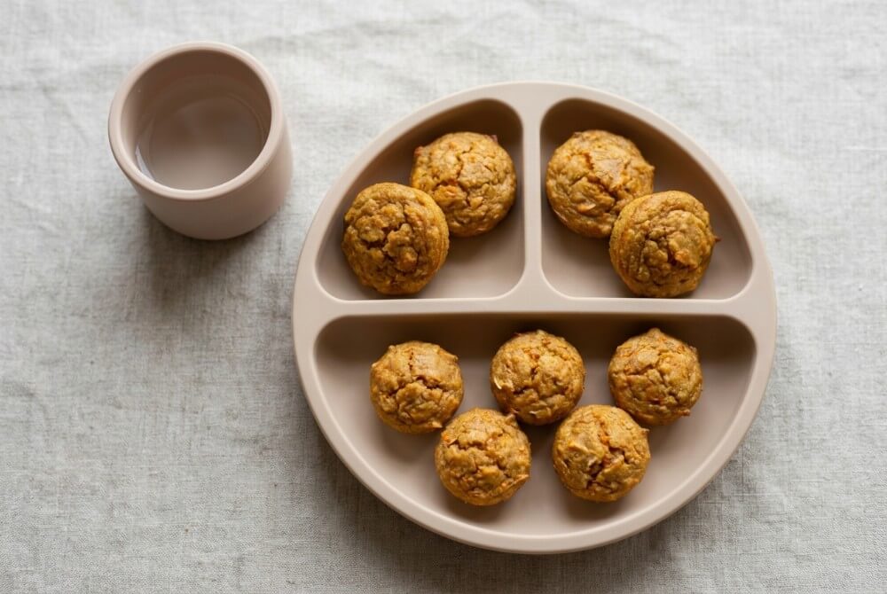 Mini muffinki marchewkowo-kokosowe dla niemowlaka ułożone na silikonowym talerzyku z przegródkami, obok mały kubeczek z wodą.