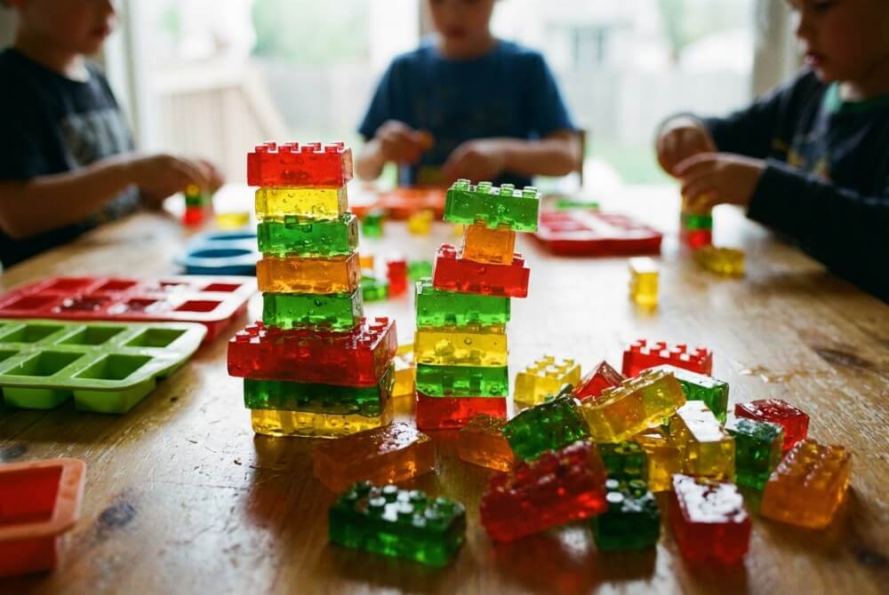 Galaretkowe klocki LEGO w intensywnych kolorach ułożone w wieżę obok silikonowych foremek na stole do zabawy.