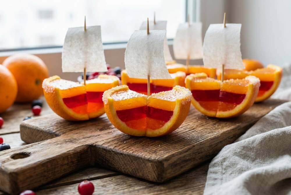 Łódeczki z galaretki w skórkach pomarańczy na drewnianej desce, część ozdobiona cukrową posypką i papierowymi żaglami.