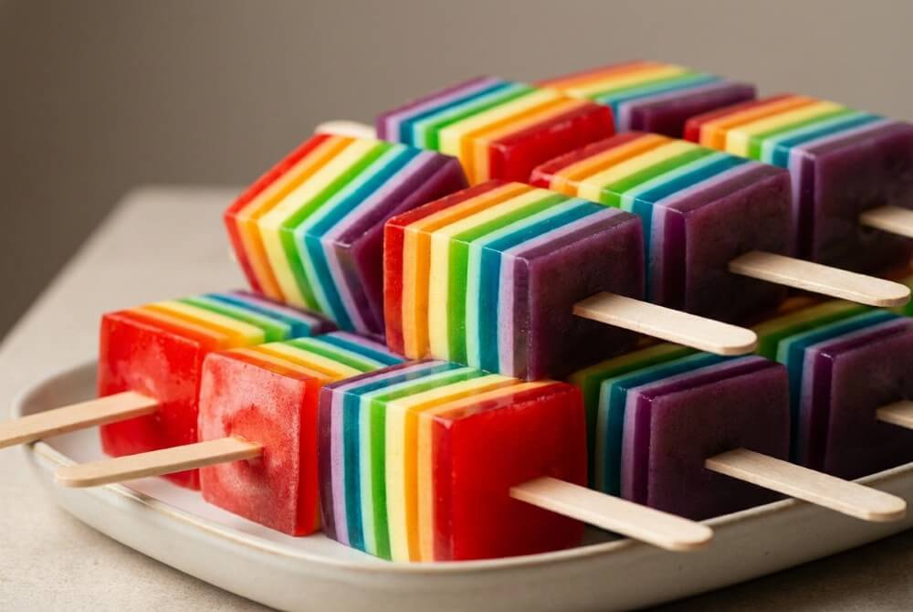 Tęczowe kostki galaretki z pianką na patyczkach, ułożone na jasnej tacy.