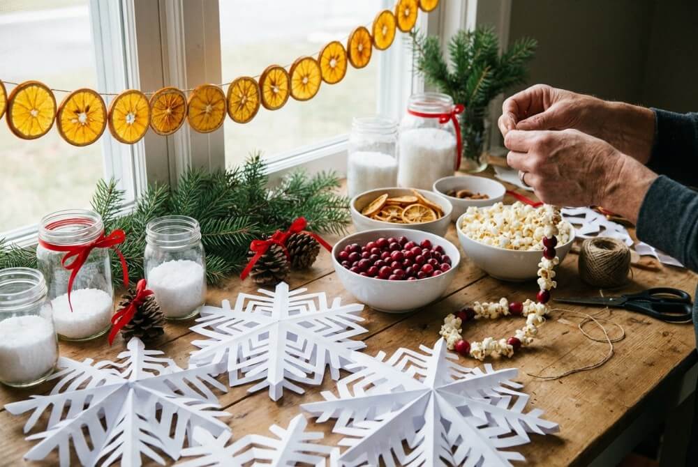 Stół z ręcznie wykonanymi świątecznymi dekoracjami DIY: suszone pomarańcze, papierowe śnieżynki 3D, stroiki w słoikach i łańcuch z popcornu i żurawiny.
