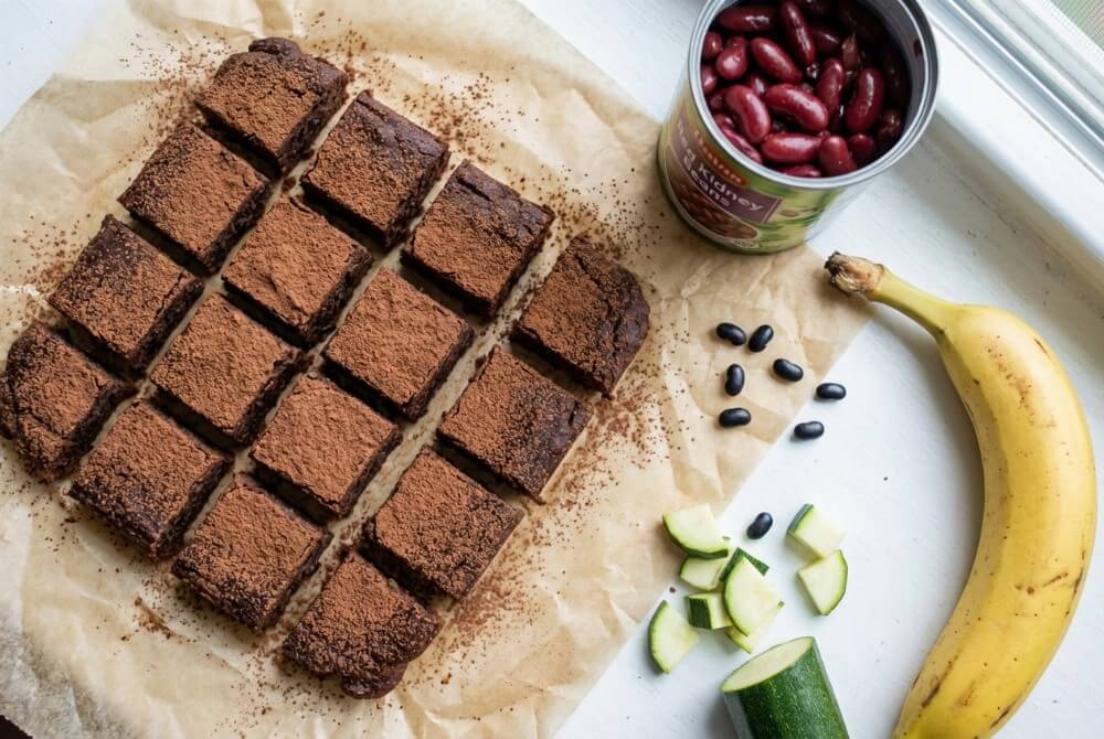 Kwadraty brownie bez mąki z fasoli (lub cukinii), posypane kakao, obok banan i fasola jako składniki.