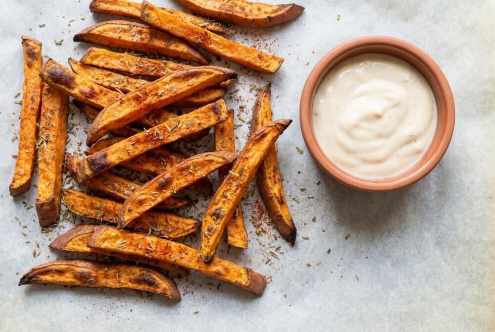 Pieczone frytki z batatów posypane ziołami prowansalskimi na blasze z papierem do pieczenia.