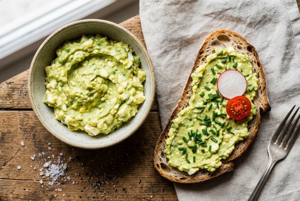 Pasta jajeczna z awokado i szczypiorkiem w miseczce oraz na kromce pieczywa.