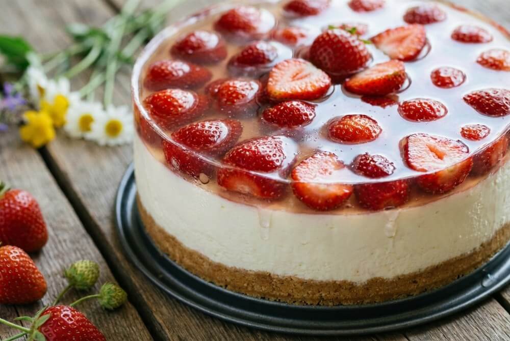 Sernik na zimno z herbatnikami, masą serową i truskawkami w galaretce w tortownicy.