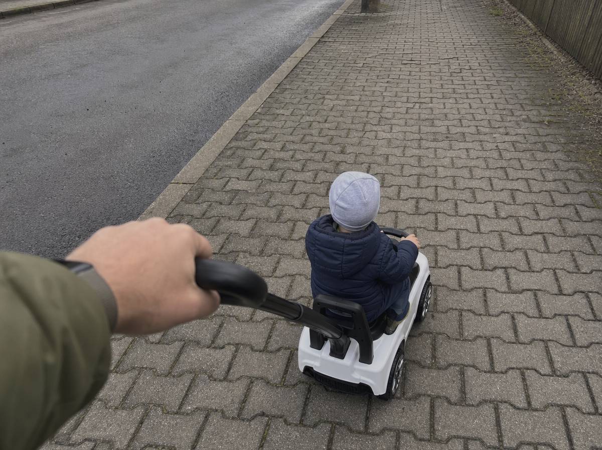 Rodzic prowadzi jeździk z rączką, dziecko siedzi z przodu na chodniku